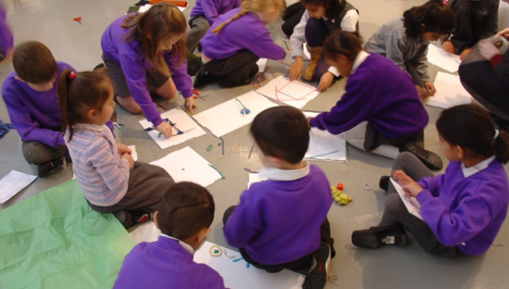 A group of school children sit on the floor using straws, pens and pencils to create drawings on their pieces of paper.