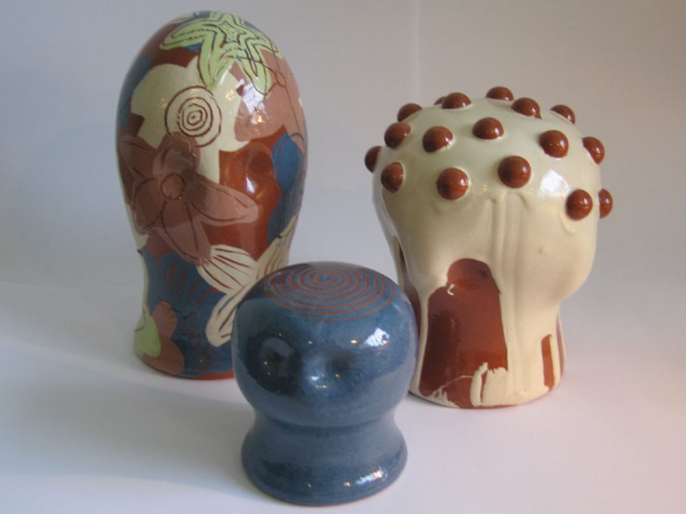 Three clay heads of various sizes with different coloured slips, shapes and decoration applied to them.
