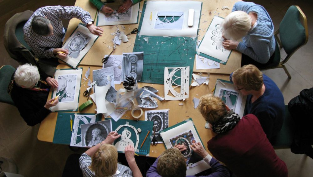 A group of people sit around a table cutting out stencils from paper.