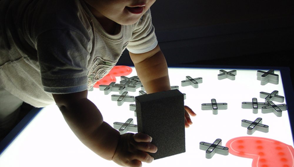 A baby leans over a lightbox, investigating plastic shapes places on top.