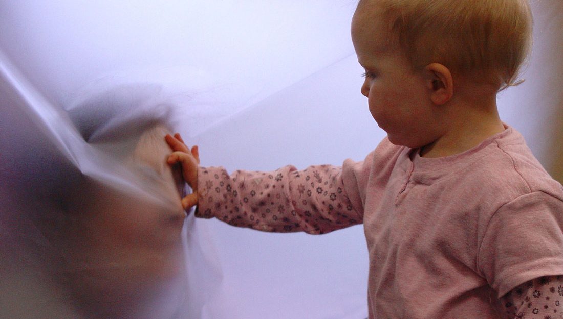 An adult pushes their face against a flat sheet of cloudy plastic. A toddler touches their face through the plastic.