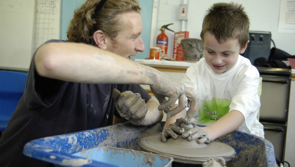 An artist helps a young boy throw a pot on the potters wheel.