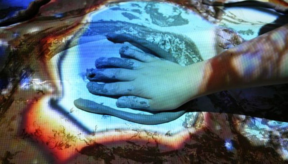 A tube of clay lies next to a hand covered in clay with images projected on to the hand.