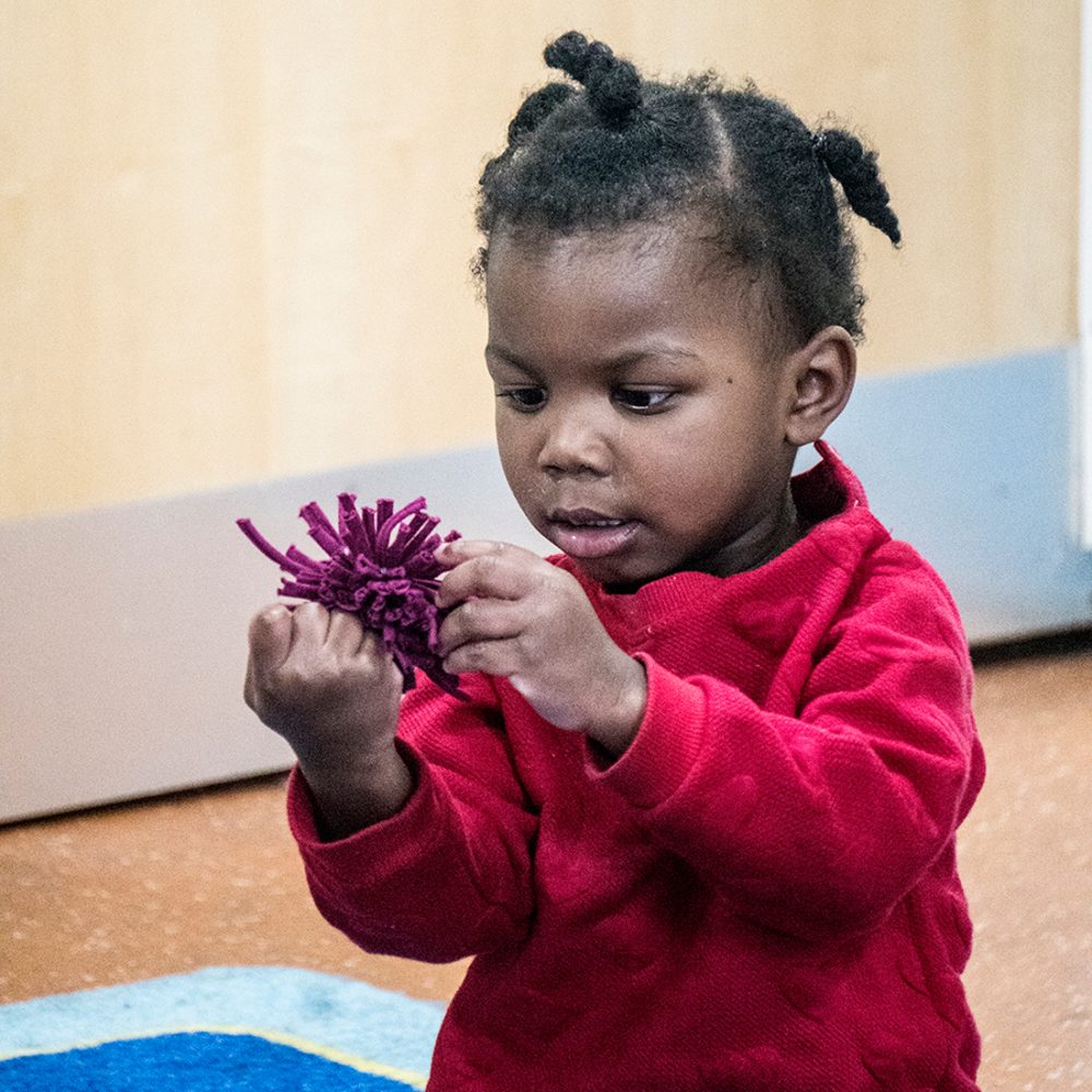 a very young girl looks closely at a pom pom and pulls on the strands of fabric.