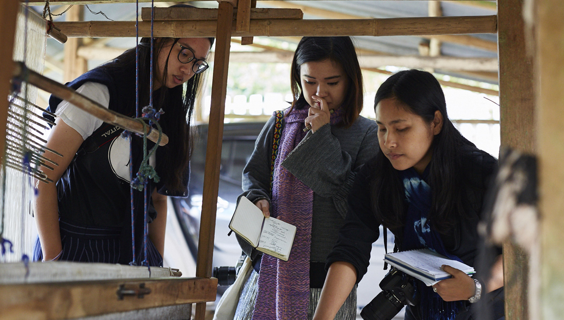 three women look at a loom, they have cameras and notebooks