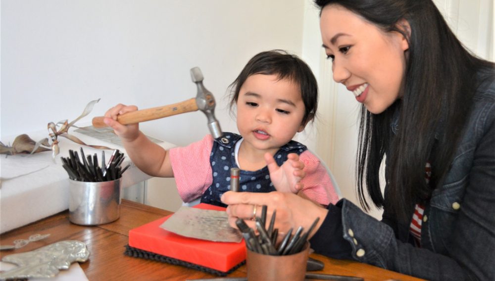 A woman and a young girl sit a a table covered in tools. The young girl, aged about 2, uses a hammer to punch some metal.