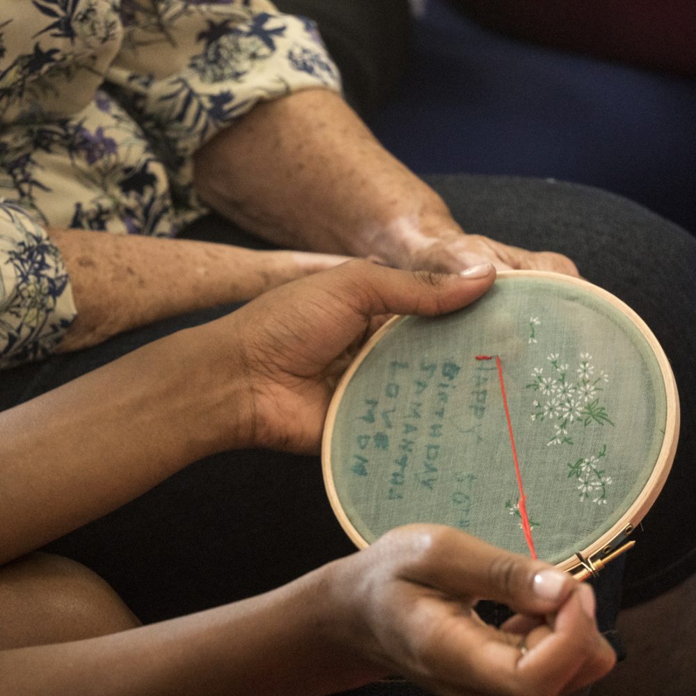 A close up of some hands embroidering simple text on material stretched over an embroidery hoop.