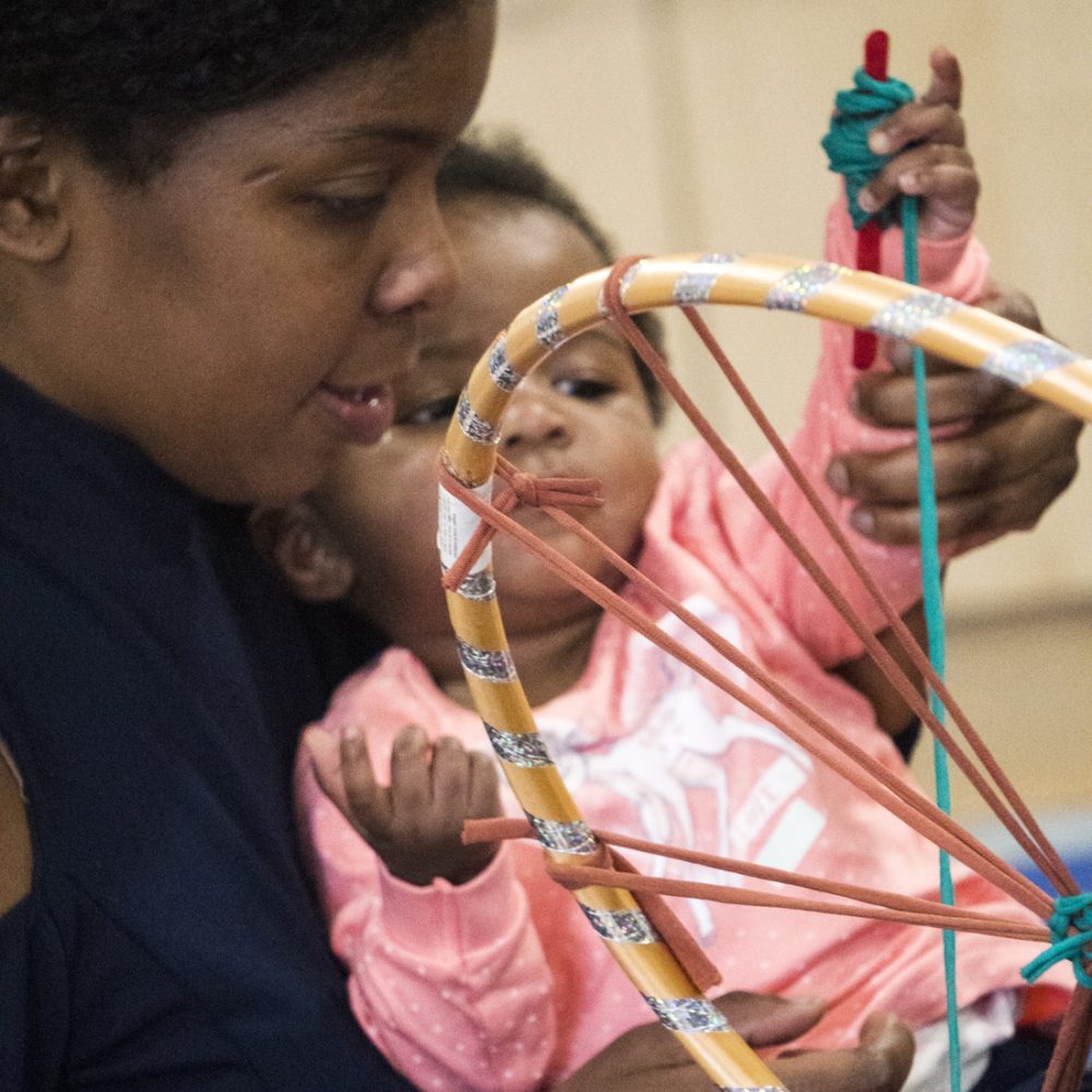 A mother and baby play with wrapping tshirt yarn around a hoop.
