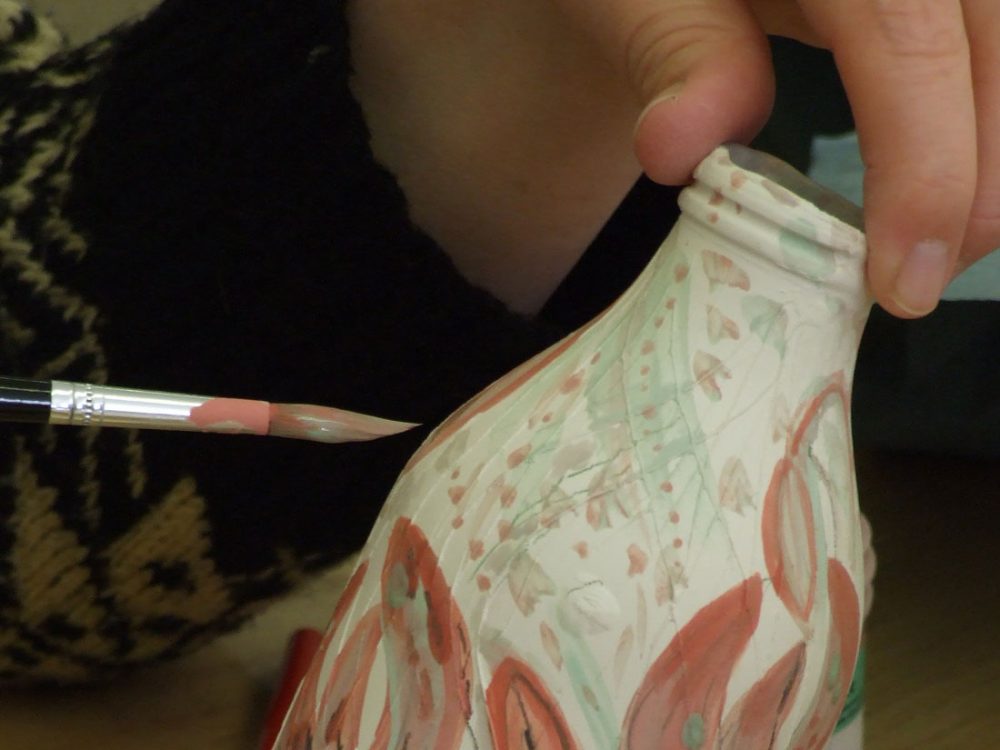 A pattern is painted onto a ceramic bottle using a paint brush.