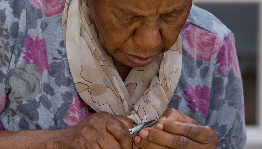 An older woman concentrates on making jewellery with some small tools.