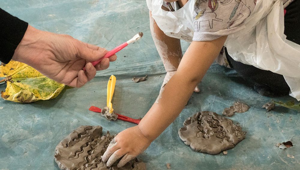 A child interacts with clay, an adult is shown passing a tool to the child.