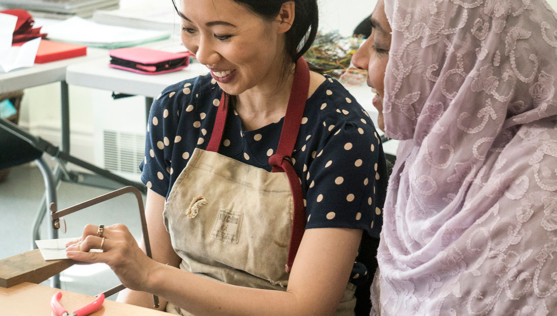 A woman shows a lady how to use a piercing saw to cut a sheet of metal. They're both smiling and happy.