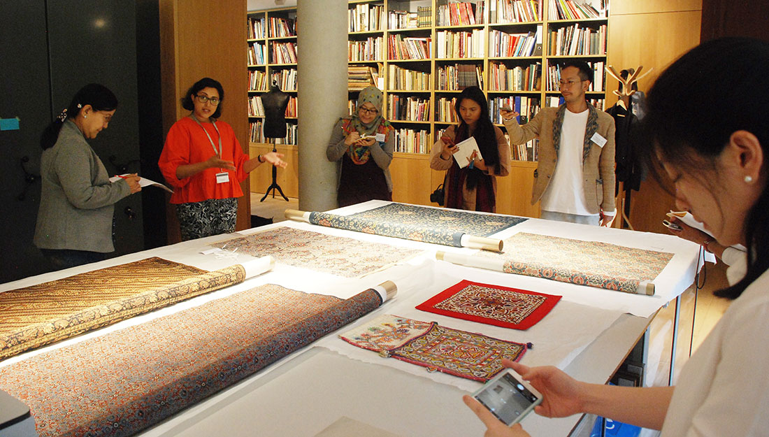 A group stand around a large table, taking notes and photos of the display of textiles.