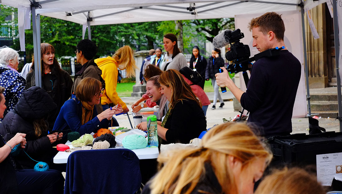 A young man films people making at a crowded table outdoors.