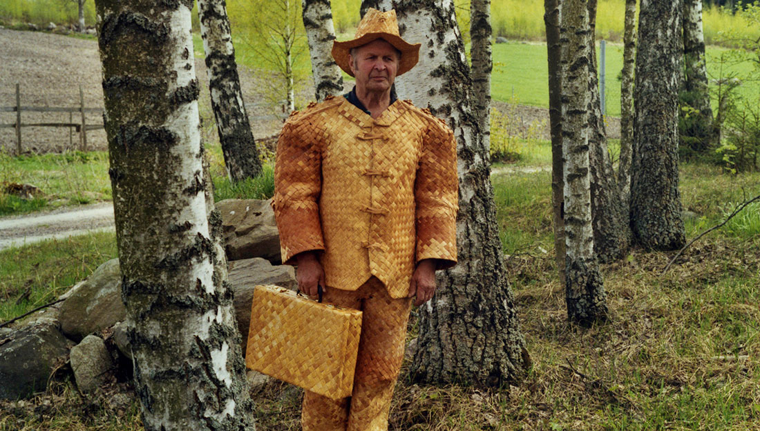 A older man, the artist, stands among birch trees dressed in a suit woven from strips of bark.