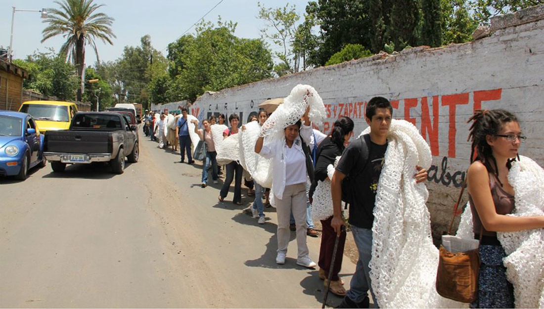 A line of people carry a very large knitted piece on their shoulders.