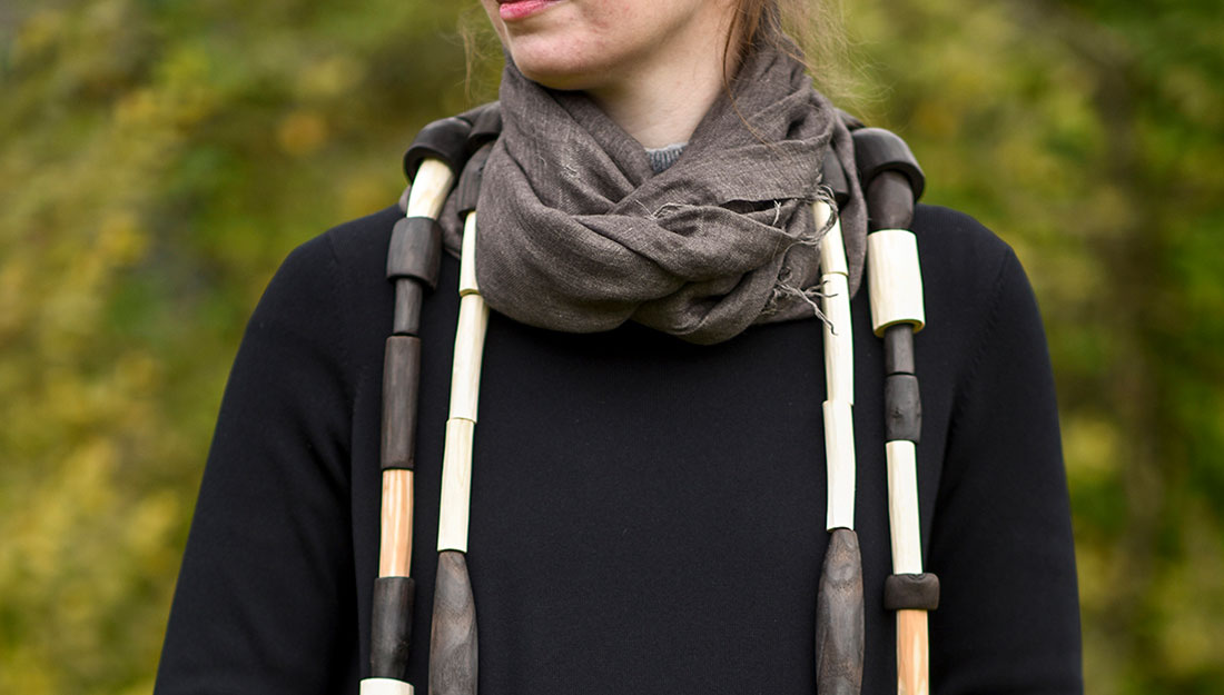 A woman wears a large neckpiece made form cylinders of wood, some are natural and some are scorched.