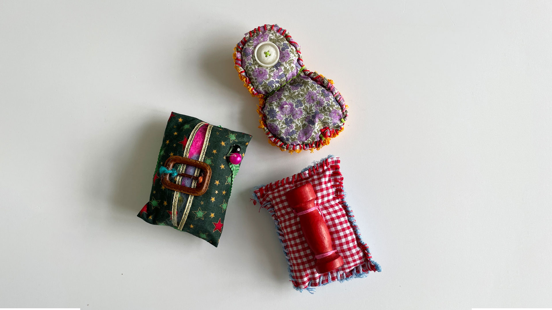 3 padded fabric shapes adorned with small objects, buttons and beads rest on a light background