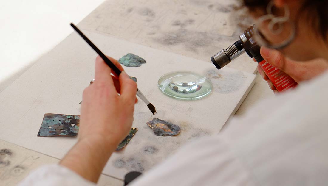 The view over a person's shoulder, holding a paintbrush in one hand and a blow torch in the other. Small cut out shapes of metal rest on a heat mat.