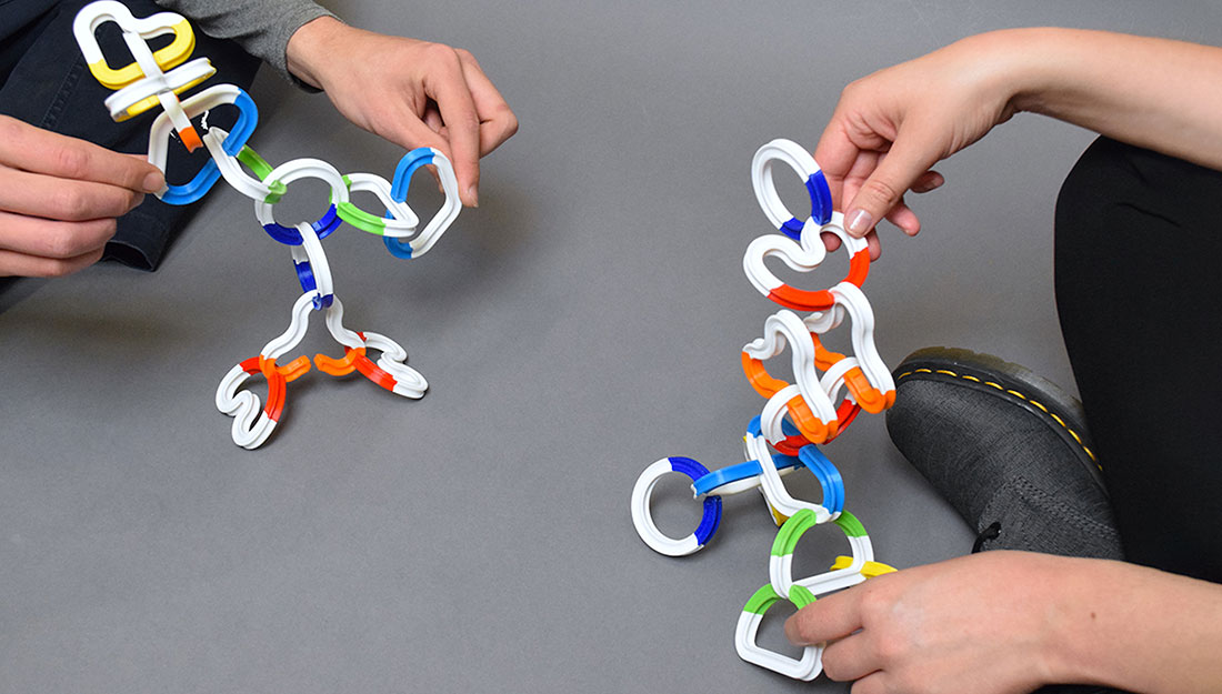 two people hold scultpures made of brightly coloured chainlike pieces.
