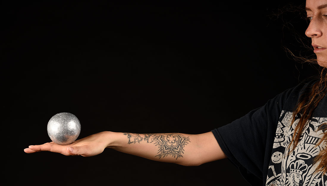 A woman holds out her arm, she balances a silver ball on her hand.