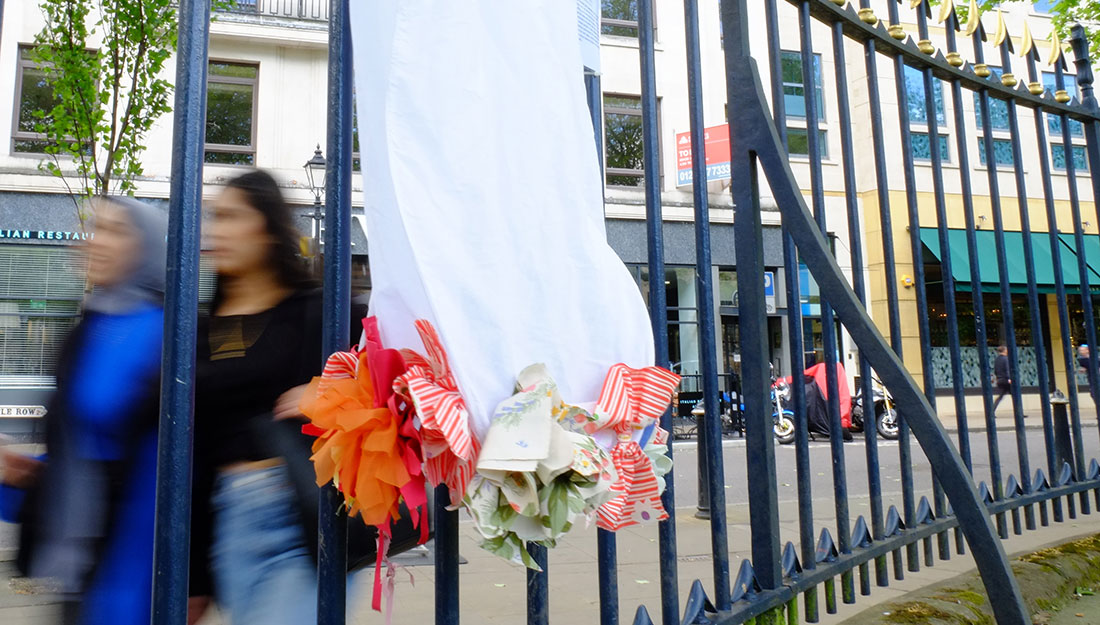 The hem of a dress decorated with fabric flower applique hangs on a railing with people walking past in the background