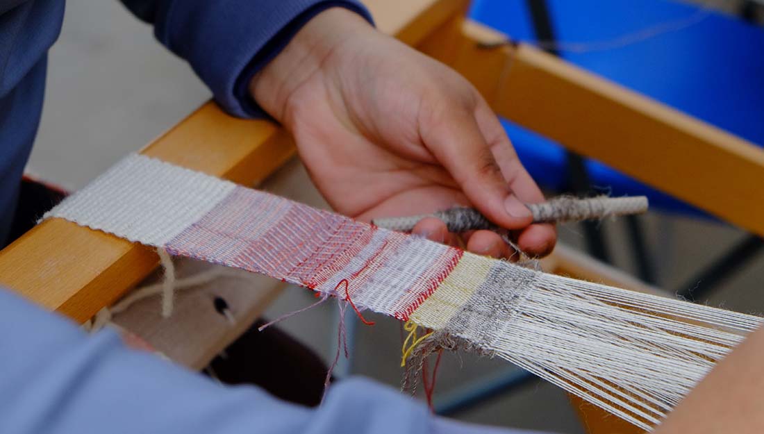 a hand holding a spool of thread working at a weaving a strip of fabric