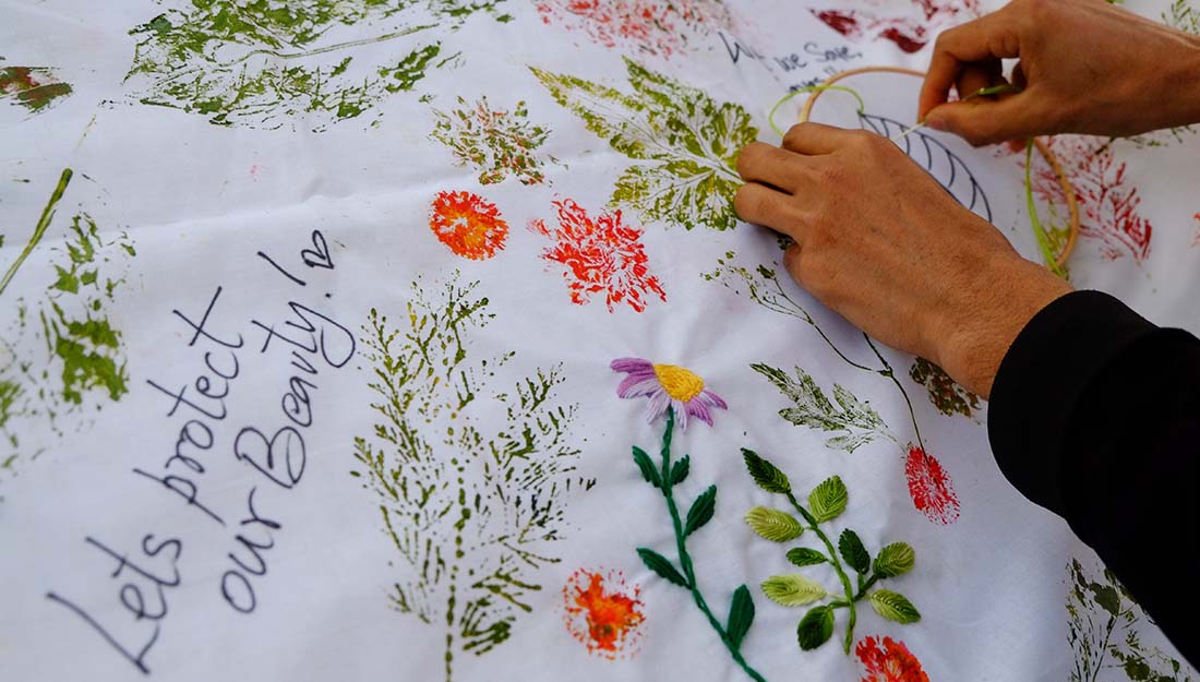A pair of hands stitching embroidery onto a large white cloth decorated with prints, text and embroidery