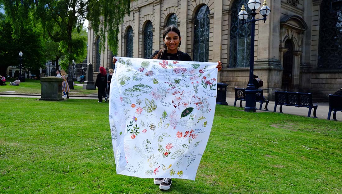 A woman stands in front of a cathedral in a park holding up a large white cloth decorated with embroidery and print