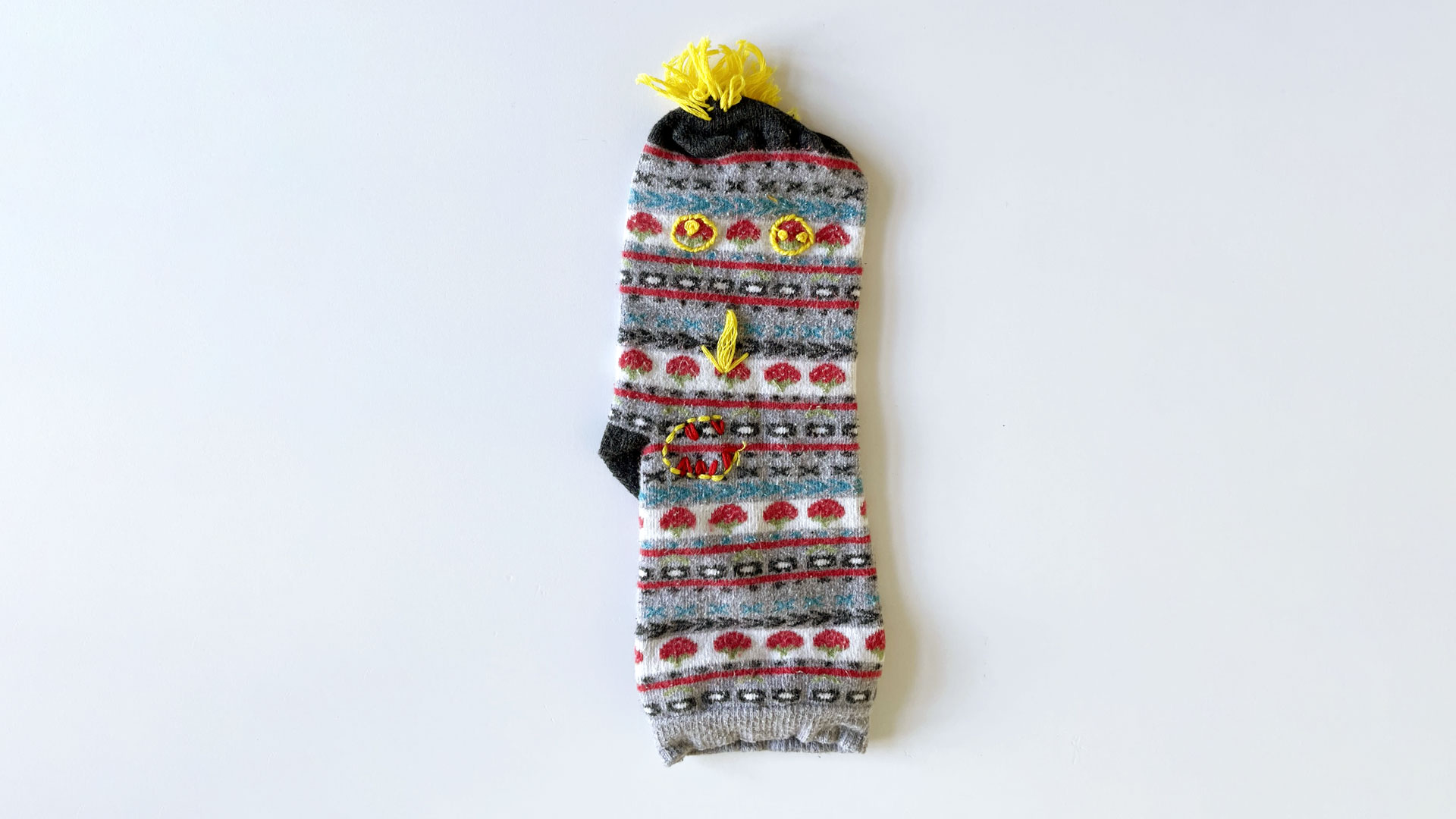 A colourful striped sock with yellow and red embroidery decoration, which resembles a face.