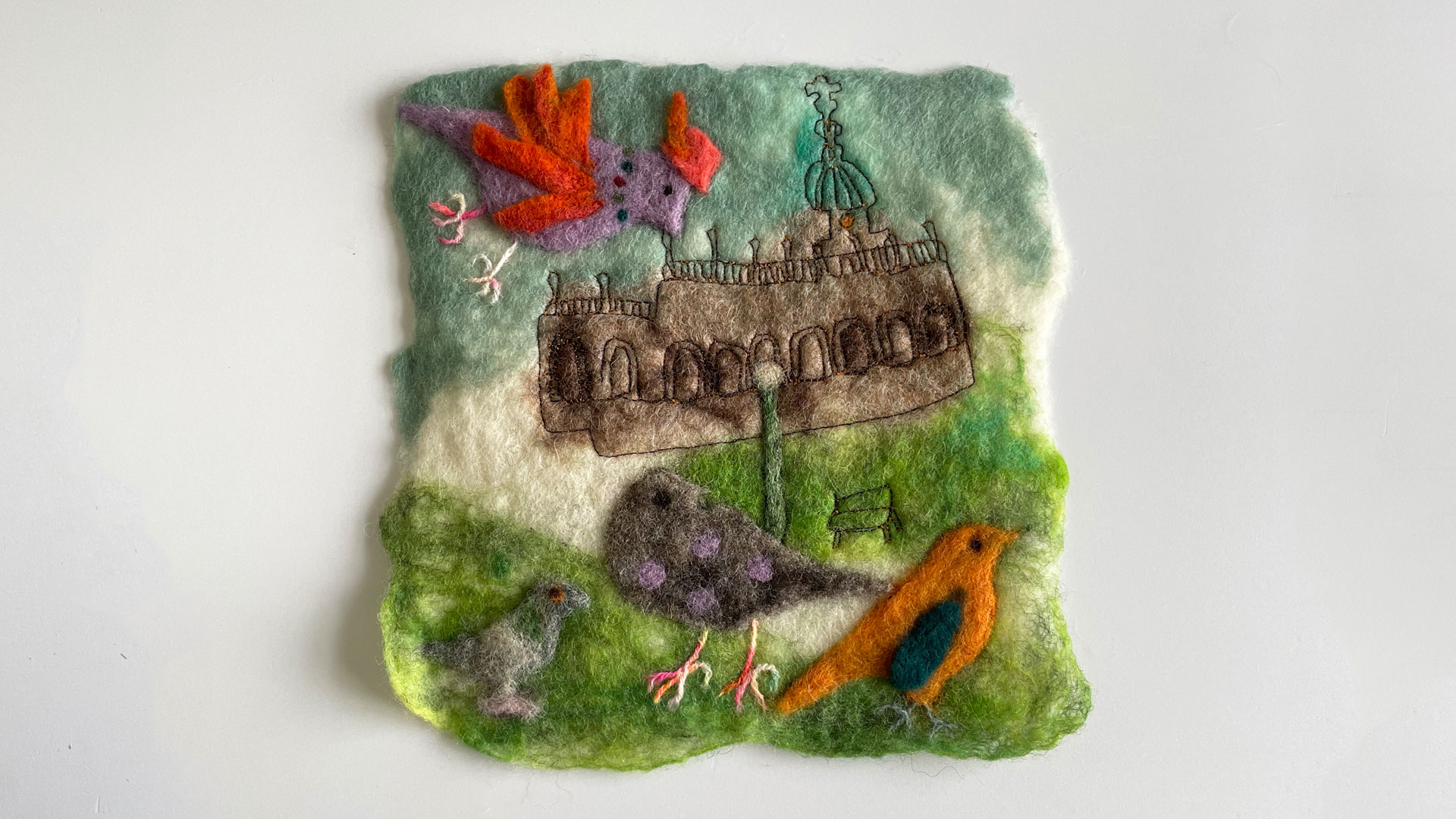 A small square felt and embroidery scene of a church and garden with 4 birds