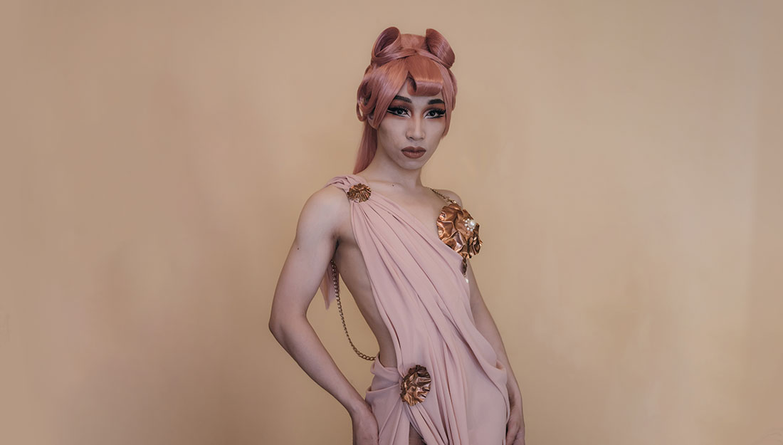 A person poses in a light pink draped and revelaing dress. The dress is adorned with flowerlike metal brooches and they are wearing striking makeup and have an unusual coral coloured wig in a glamourous style.