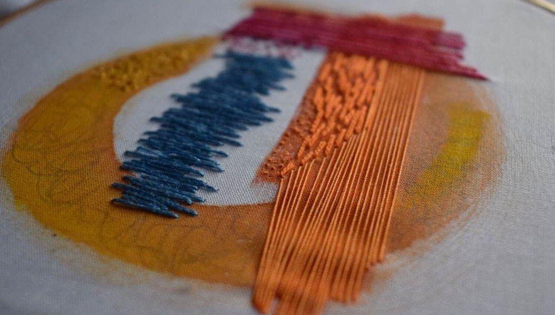A bright abstract embroidery: a yellow circle, with orange, blue nd red shapes embroidered.