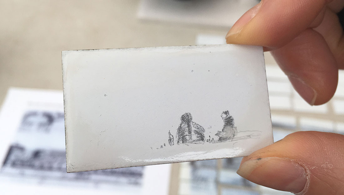 A small enamel tile with a drawing of two people sitting on the grass.