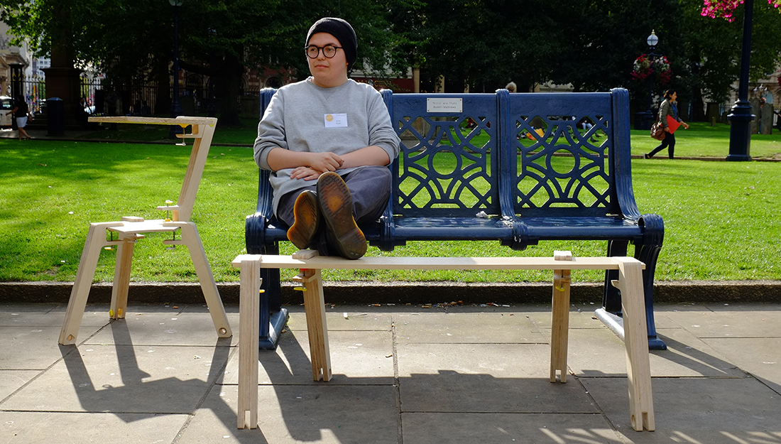 The artist sits on a bench surrounded by furniture made from discarded wood.