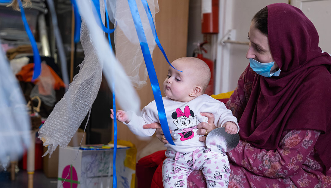 A baby sits on an adult's knee reaching for hanging strips made of various materials.