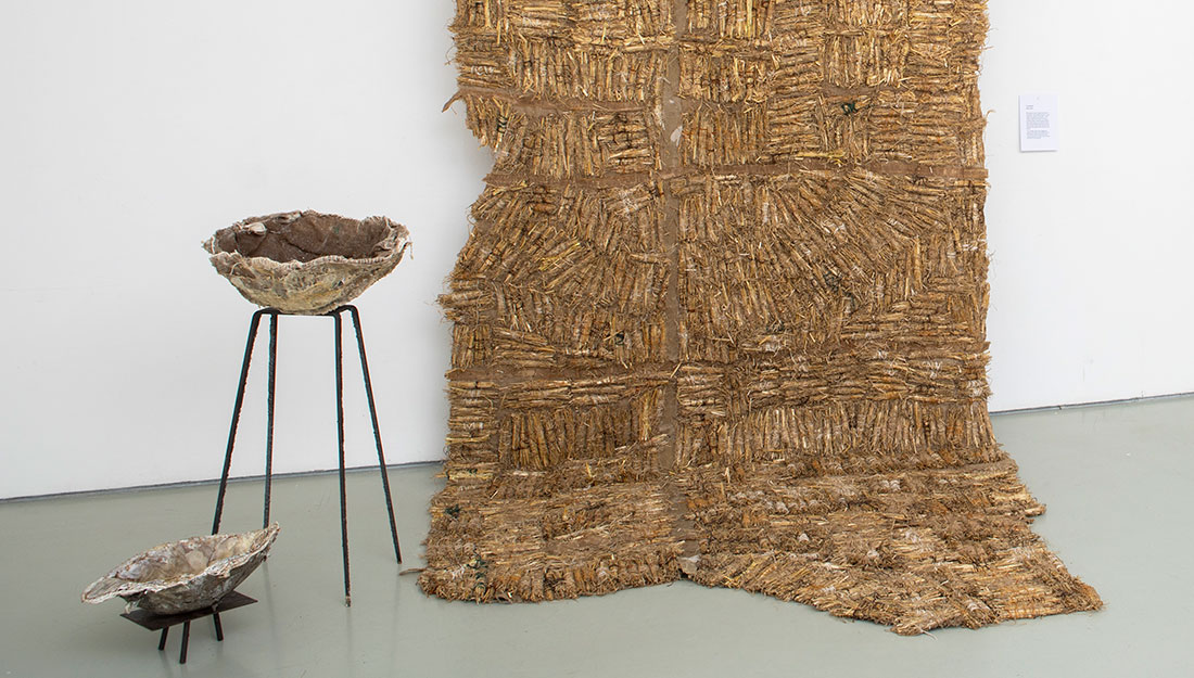 A close up of the exhibit, two rustic bowls on black stands and a large hanging decorated with small bundles of straw.