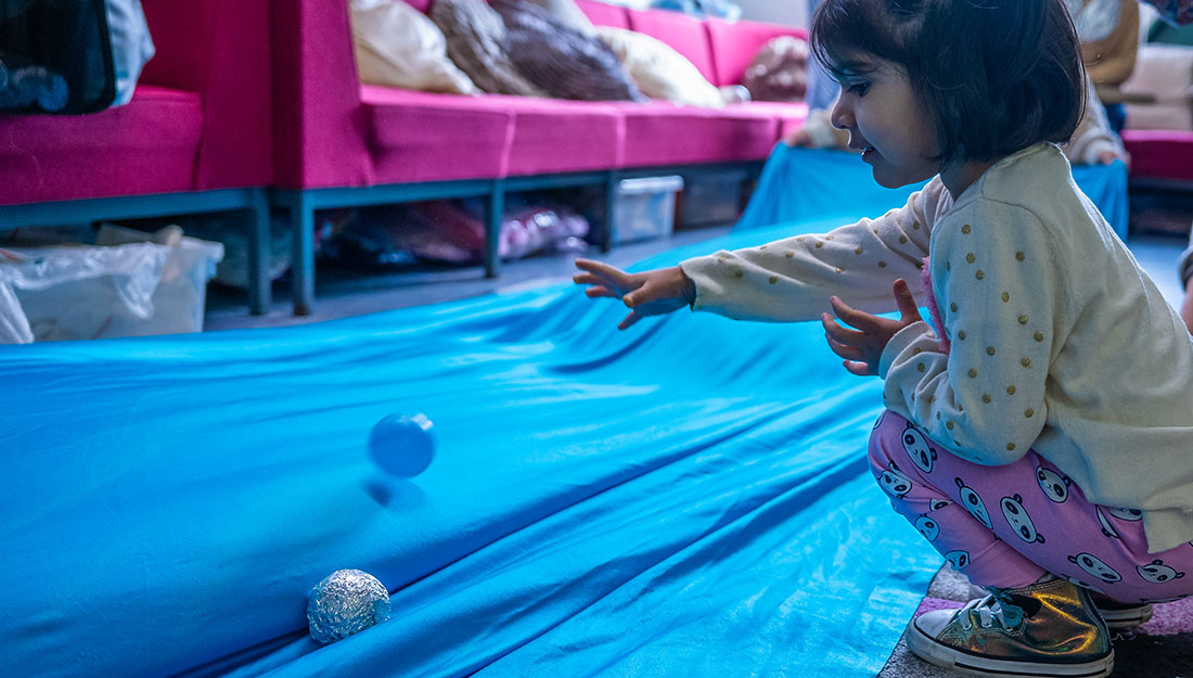 A smiling toddler throws plastic and foil covered balls onto a very large bright blue fabric sheet.