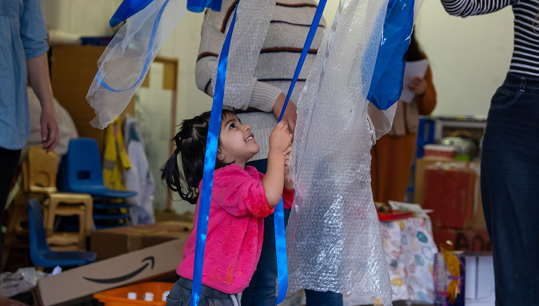 A smiling toddler looks up and reaches for strips of ribbon and material.