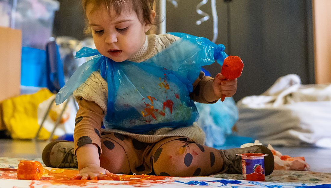 A toddler concentrates as she spreads paint onto paper with her hand. She is messy and calm.