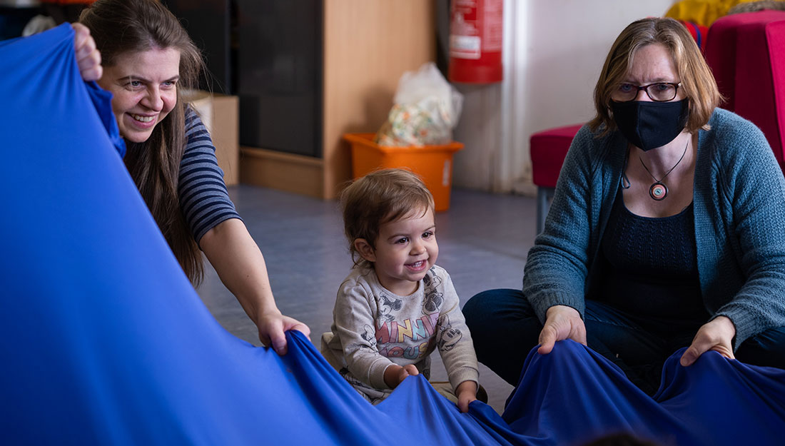 smiling adults and a toddler hold and move the edge of a very large piece of blue fabric.