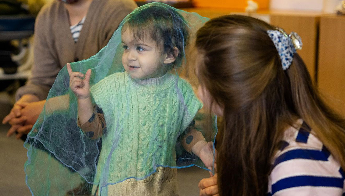 A smiling toddler is draped a very thin mesh material. An adult watches them.