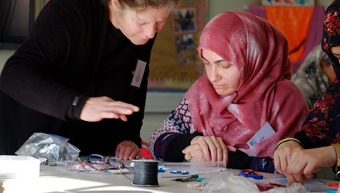 A woman is teaching jewellery skills to another women who is wearing a pink headscarf.