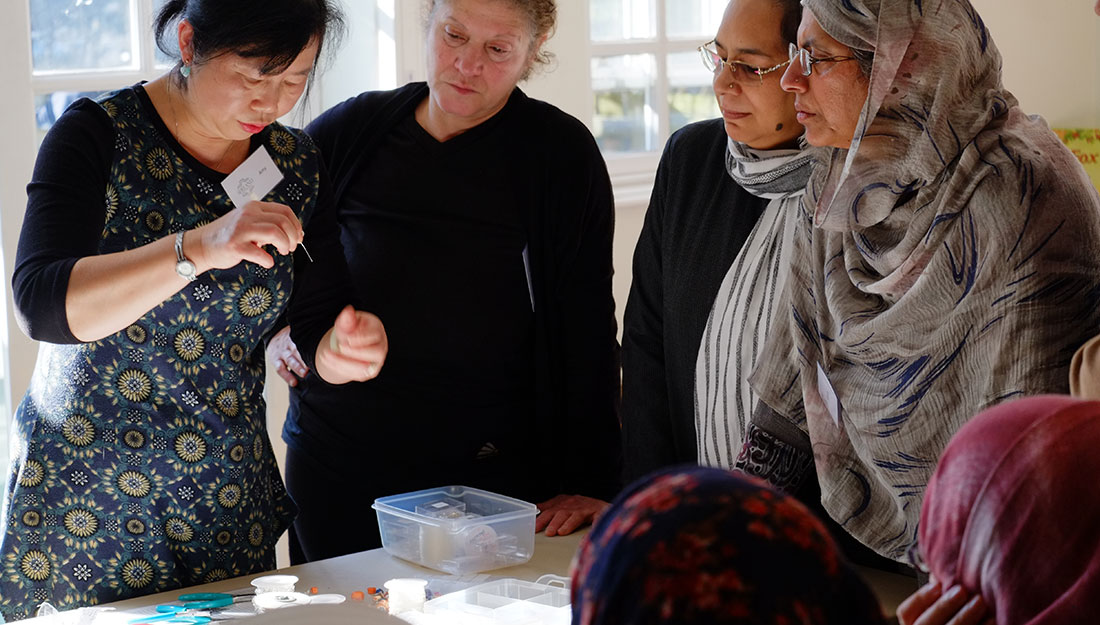 Four women of different cultures stand around a table. One person is demonstrating a jewellery technique.