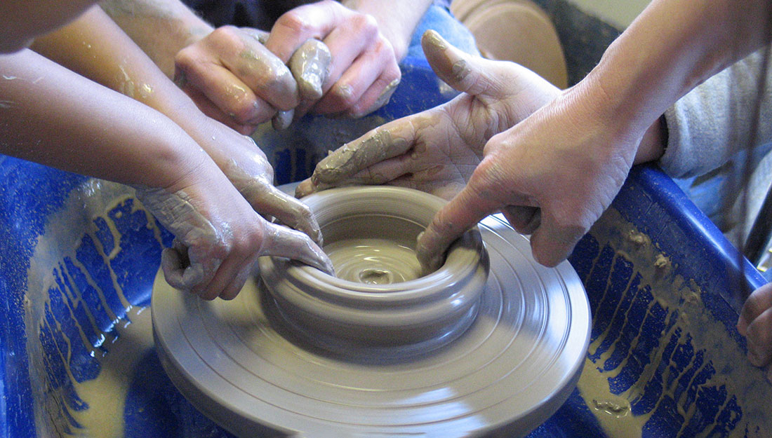 Adult and very young hangs reach for clay spinning on a potters wheel.
