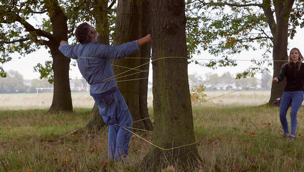 A man wearing a denim shirt and jeans in a park leaning on yellow strings which are tied around a tree. A woman with brown hair is opposite the man in the park, holding onto one of the strings to support the man leaning.