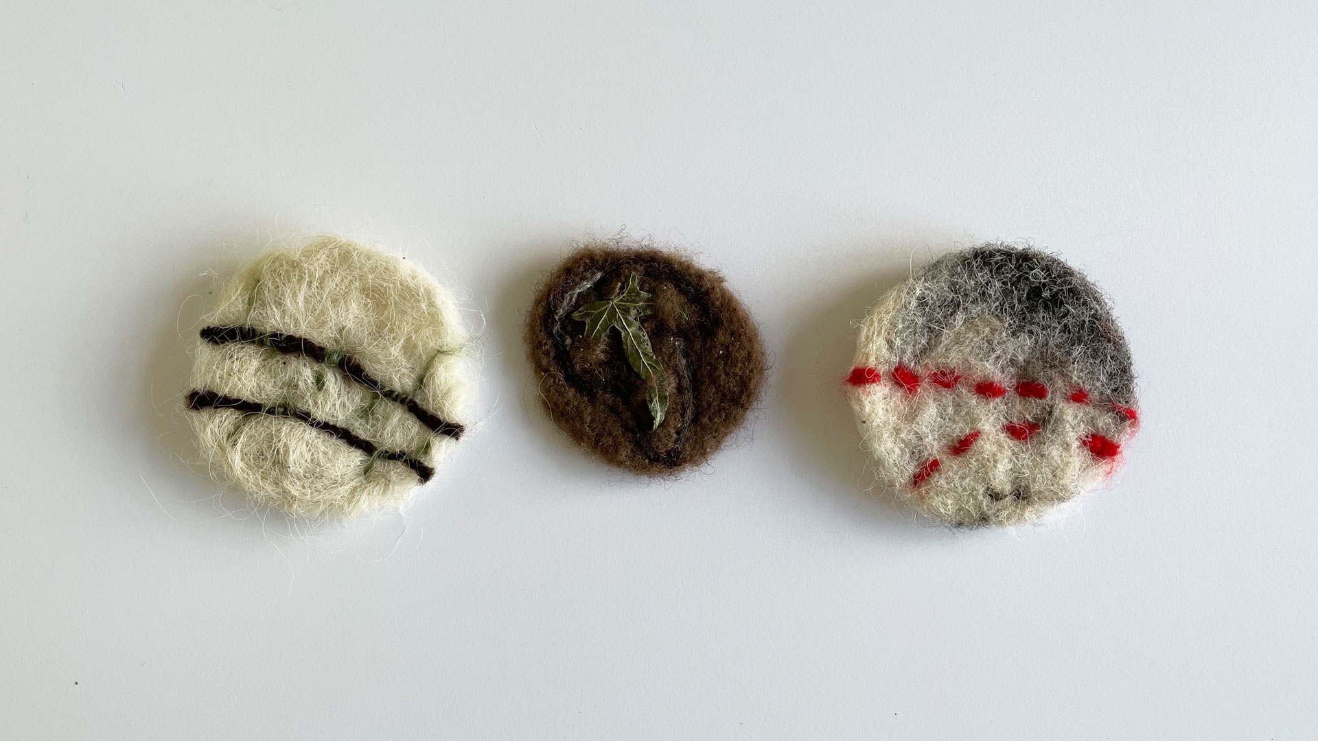 3 circular brooches made of felted wool and natural materials on a white background