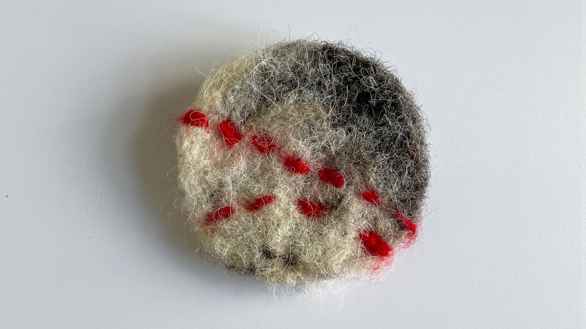 A circular brooch made of a mixture of brown and natural coloured wool with two red stripes, on a white background