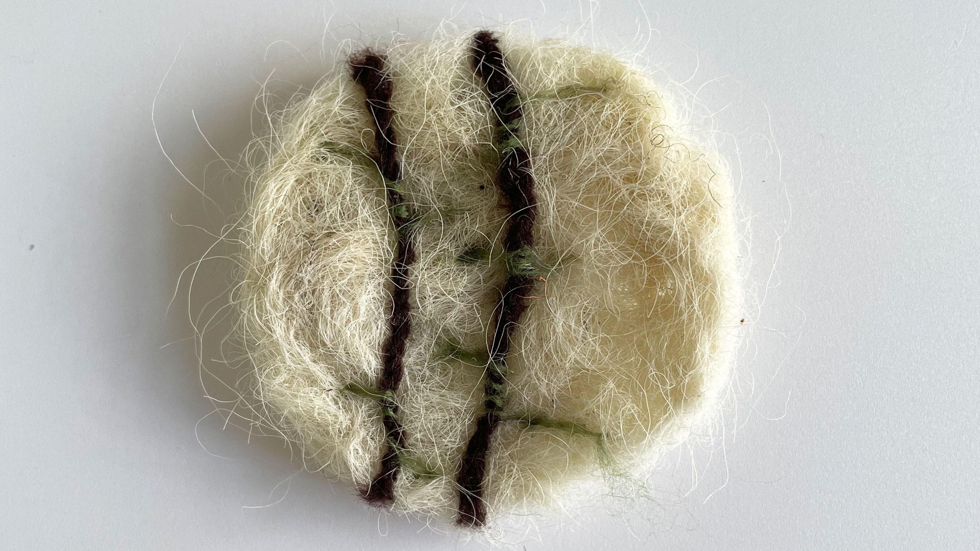 A circular brooch made of white felted wool with two brown stripes with green decoration, on a white background