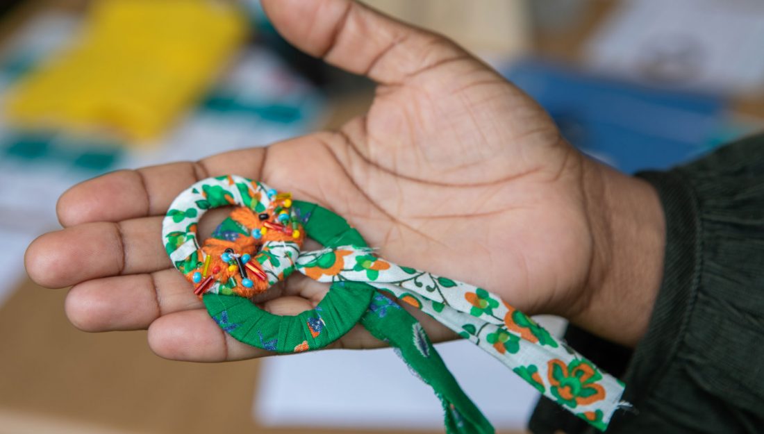 a hand holding a handmade textile object made up of two rings wrapped in green, orange and white yarn